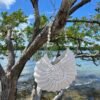 White crocheted purse hanging from driftwood branch against ocean backdrop with rocky shore and blue sky - angle 2