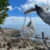 White crocheted purse hanging from driftwood against coastal ocean view with rocky shore, angle 1