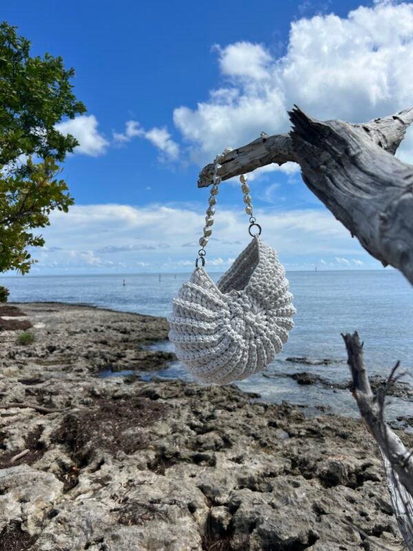 White crocheted purse hanging from driftwood against coastal ocean view with rocky shore, angle 1